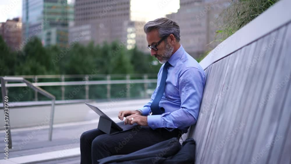 Successful Mature businessman in blue shirt and suit sitting on bench while typing text for email on touch pad. Caucasian entrepreneur in stylish eye glasses using tablet for work and answering call Successful Mature businessman in blue shirt and suit sitting on bench while typing text for email on touch pad. Caucasian entrepreneur in stylish eye glasses using tablet for work and answering call