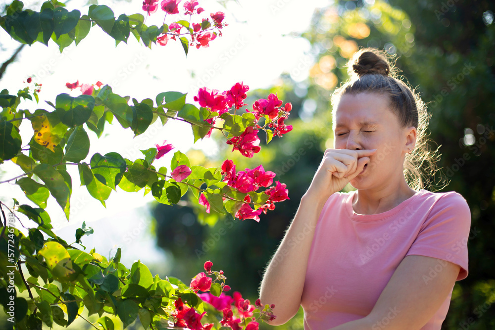Portrait of beautiful young allergic woman is suffering from pollen allergy or cold on natural flower flowering tree background at spring or sunny summer day sneezes, blowing her runny nose rubs eyes