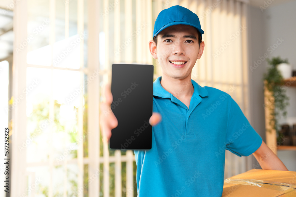 Asian delivery man holding mobile mockup template design with cardboard box for express shipping while wearing blue uniform and cap at the home office Asian delivery man holding mobile mockup template design with cardboard box for express shipping while wearing blue uniform and cap at the home office