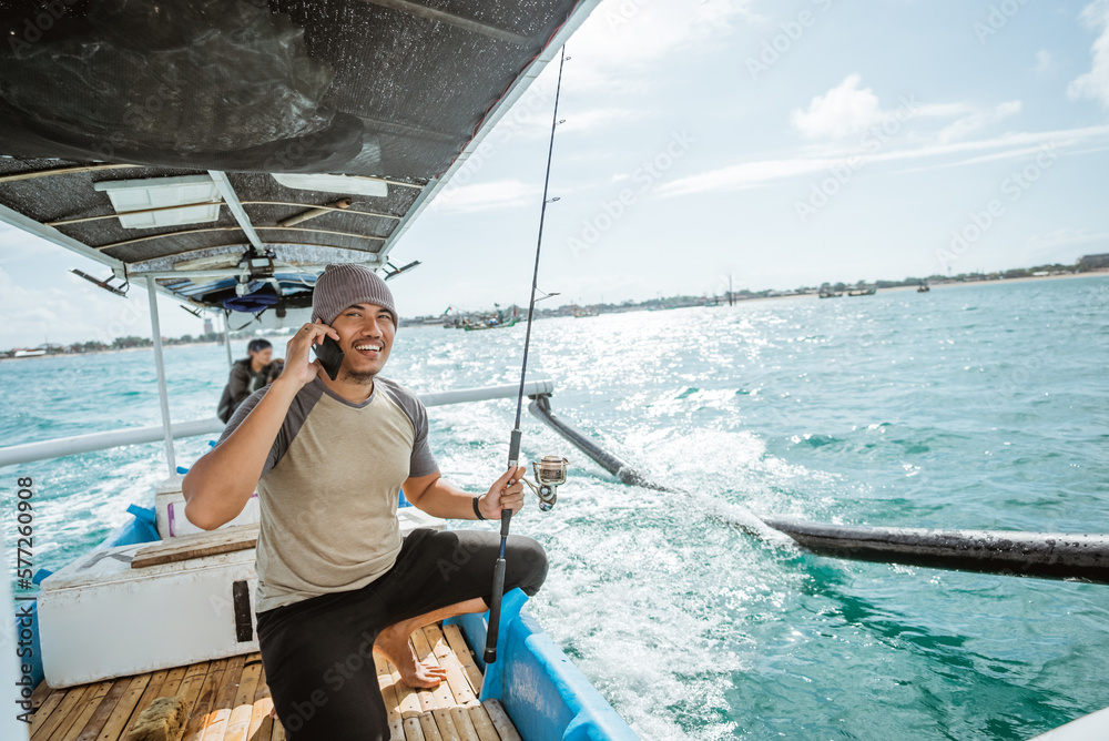 asian fisherman making a cell phone call while holding a fishing rod on a small fishing boat asian fisherman making a cell phone call while holding a fishing rod on a small fishing boat