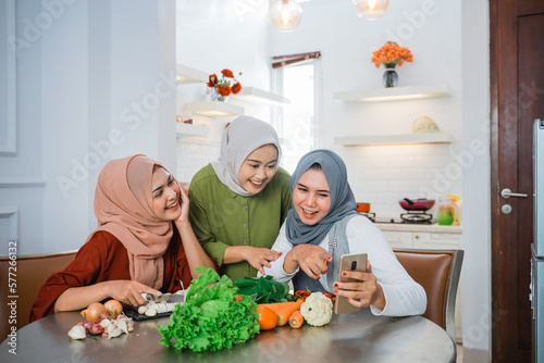 happy young asian woman with hijab cooking in the kitchen while looking at internet on mobile phone for menu today