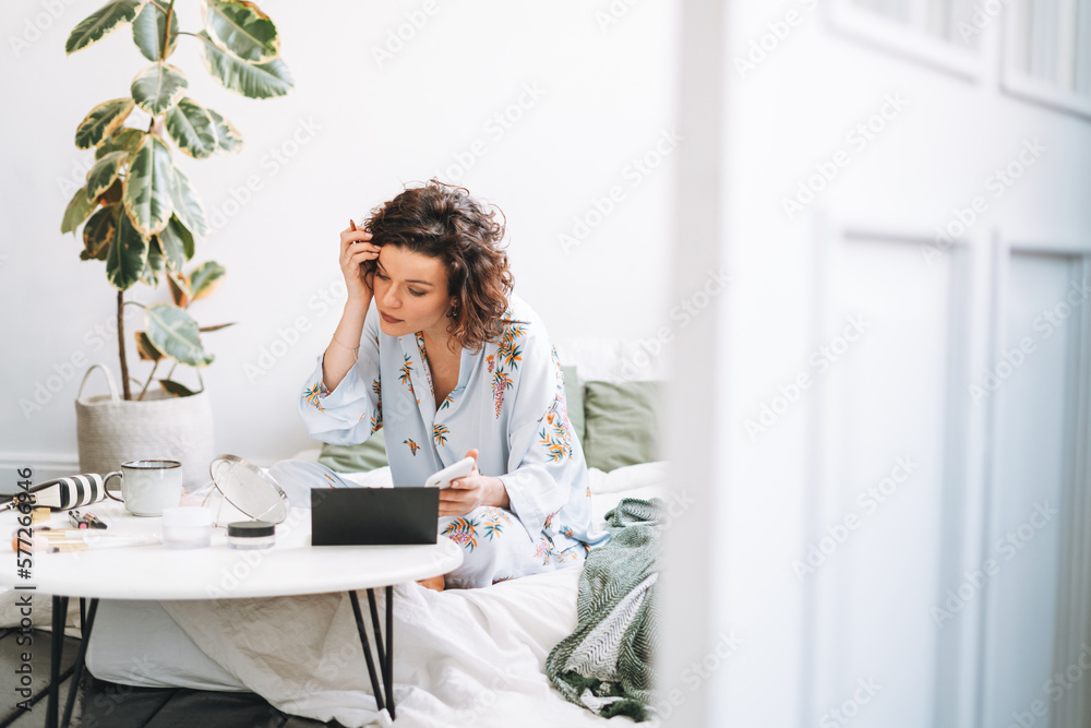 © Galina Zhigalova - Young brunette woman in blue pajamas applies makeup with mobile phone sitting on bed at home © Galina Zhigalova - Young brunette woman in blue pajamas applies makeup with mobile phone sitting on bed at home