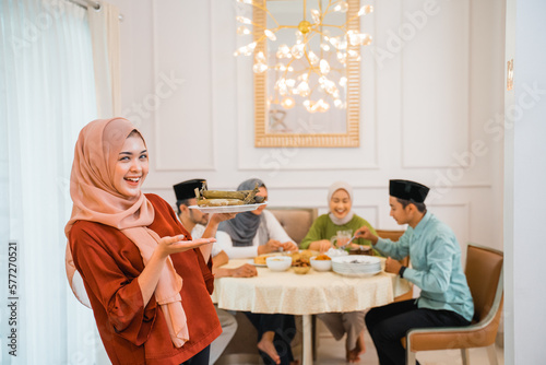 Beautiful veiled girl carrying a plate of lontong rice cake. muslim family together enjoy the iftar meal in the dining room.