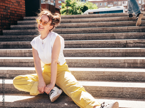Young beautiful smiling hipster woman in trendy summer yellow jeans clothes. Carefree teen model posing in the street at sunset. Positive female outdoors. Sitting at the stairs. In sunglasses © halayalex