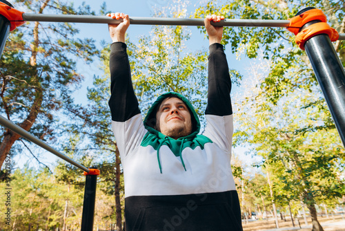Male man hanging on bars on sunny autumn day. Outdoor park sports equipment. Lifestyle portrait of young caucasian unshaven man 34 years old in hooded hoodie.
