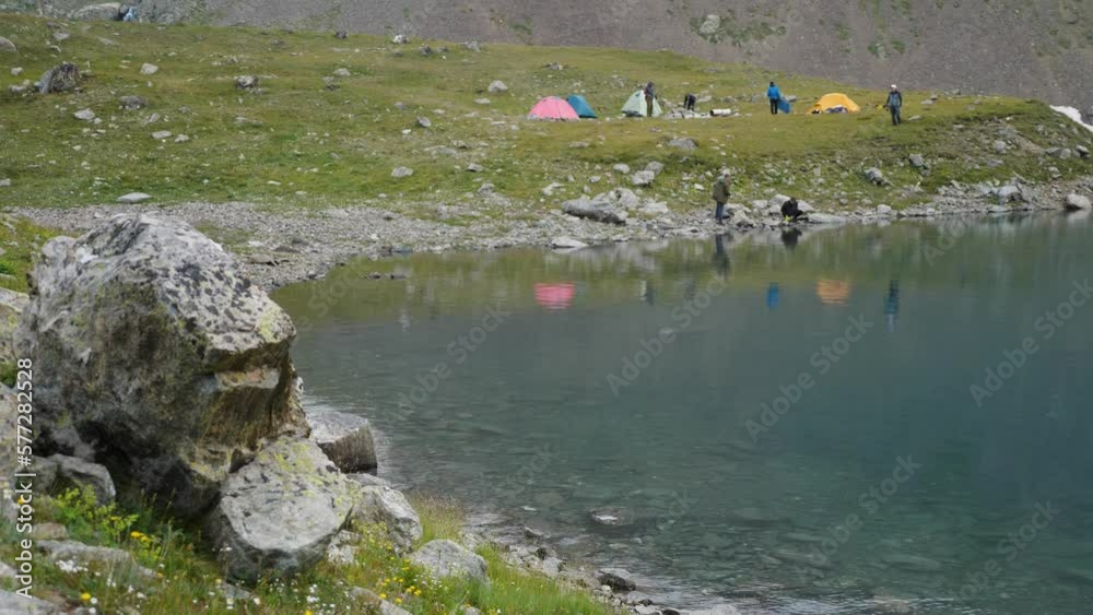 A serene blue mountain lake with crystal clear water. Against backdrop of mountain. On the shore of the lake is a tourist camp. Colourful tents dot the landscape, and tourists walk around leisurely A serene blue mountain lake with crystal clear water. Against backdrop of mountain. On the shore of the lake is a tourist camp. Colourful tents dot the landscape, and tourists walk around leisurely