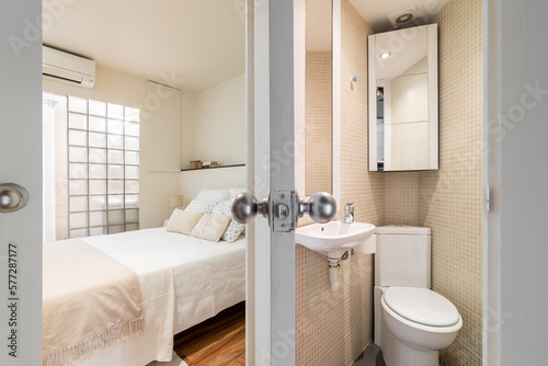 View from bathroom with toilet with beige tiles toilet bowl and sink to bedroom with bed and glass brick wall. Concept of cramped but thoughtful interiors and space