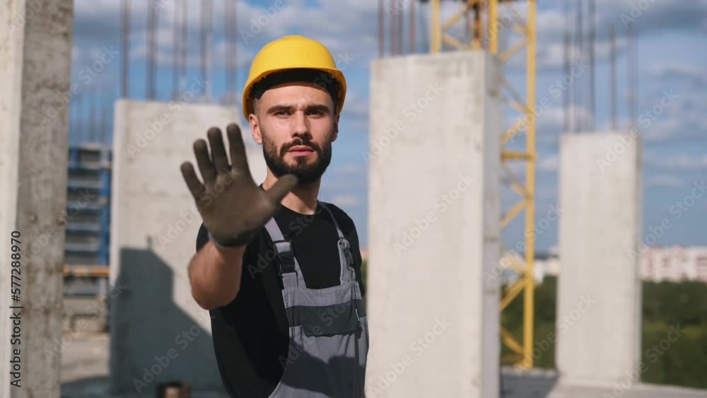 Showing stop gesture by hand. Young engineer in hard hat and uniform is on the construction site. Showing stop gesture by hand. Young engineer in hard hat and uniform is on the construction site.