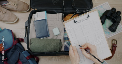 Close-up of travel checklist and packed suitcase and female hand checking items getting ready for trip. Tourism and baggage preparation concept.