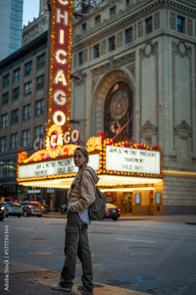 Fototapeta premium Portrait of a long-haired blonde young girl with a sweatshirt, backpack and vintage photo camera sightseeing at night in Chicago, in front of the famous theater