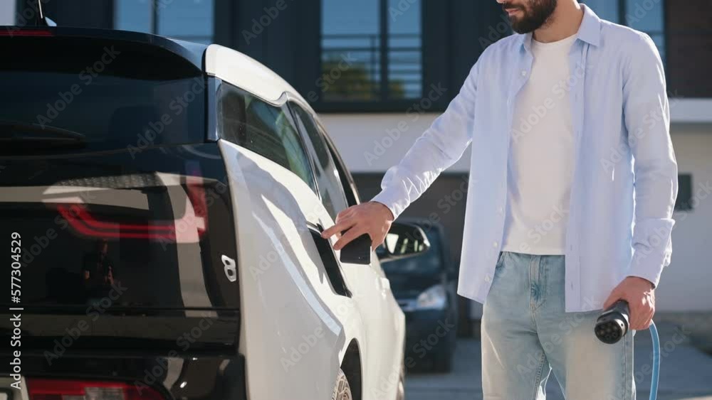 Walking and putting charging cable into vehicle. Stylish young man in white clothes is outdoors. Walking and putting charging cable into vehicle. Stylish young man in white clothes is outdoors.