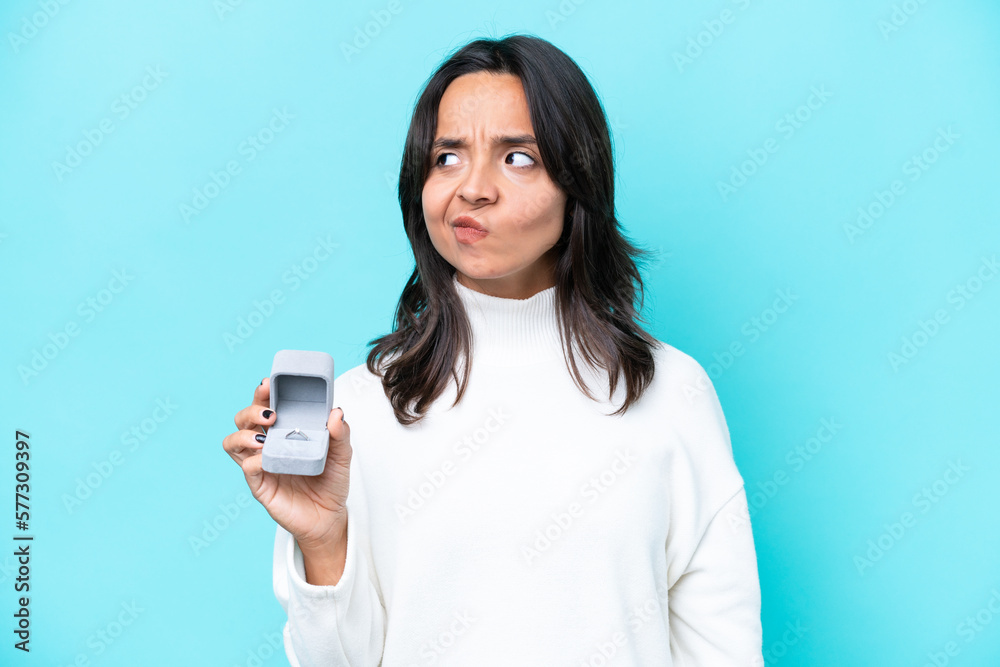 Young hispanic woman holding a engagement ring isolated on blue background having doubts and thinking Young hispanic woman holding a engagement ring isolated on blue background having doubts and thinking