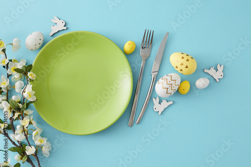 Easter concept. Top view composition of green plate colorful easter eggs cutlery cute rabbits and cherry blossom branch on isolated pastel blue background with blank space