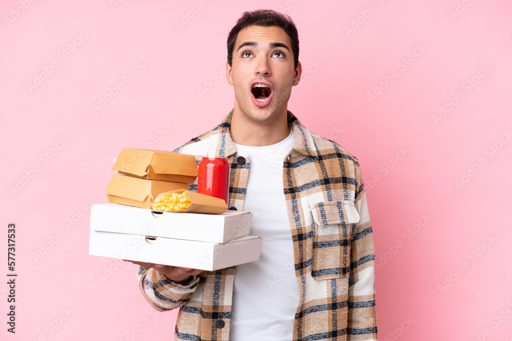 Young caucasian man holding fast food isolated on pink background looking up and with surprised expression Young caucasian man holding fast food isolated on pink background looking up and with surprised expression