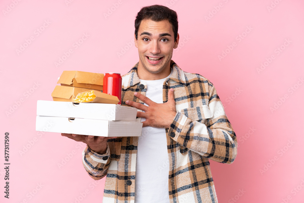 Young caucasian man holding fast food isolated on pink background surprised and shocked while looking right Young caucasian man holding fast food isolated on pink background surprised and shocked while looking right