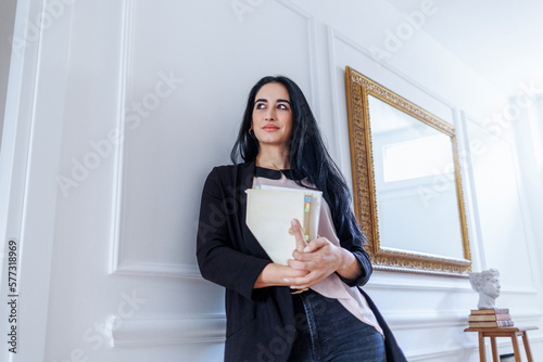 donna mora con i capelli neri sta in piedi tenendo dei libri in mano appoggiata nel salotto di casa sua