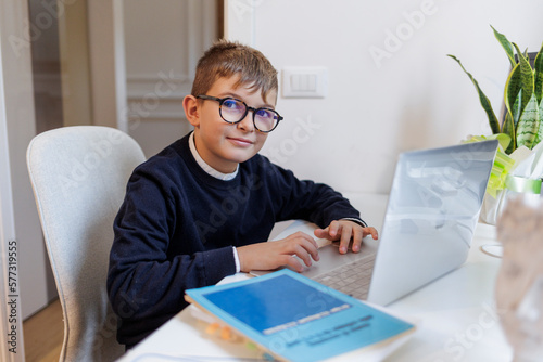 Bambino biondo con gli occhiali , vestito in blu e seduto davanti a una scrivania mentre lavora al computer photo