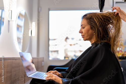 Side view of adult focused female in cape using netbook working remotely while crop hairdresser drying long hair