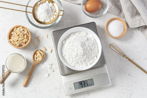 Flour in white bowl measuring on digital scale with cake or bakery ingredients and utensil on marble kitchen table
