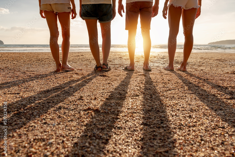 Group of friends stands at sunset sea beach. Closeup photo of legs and long shadows - obrazy, fototapety, plakaty 