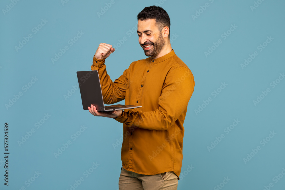 Excited italian man holding laptop pc computer doing winner gesture clench fist isolated on blue background Excited italian man holding laptop pc computer doing winner gesture clench fist isolated on blue background