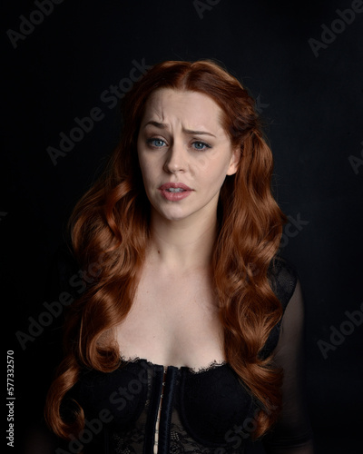 close up portrait of beautiful woman with long red hair wearing sheer corset top. variety of facial expressions,  Isolated on dark studio background with moody silhouette lighting.