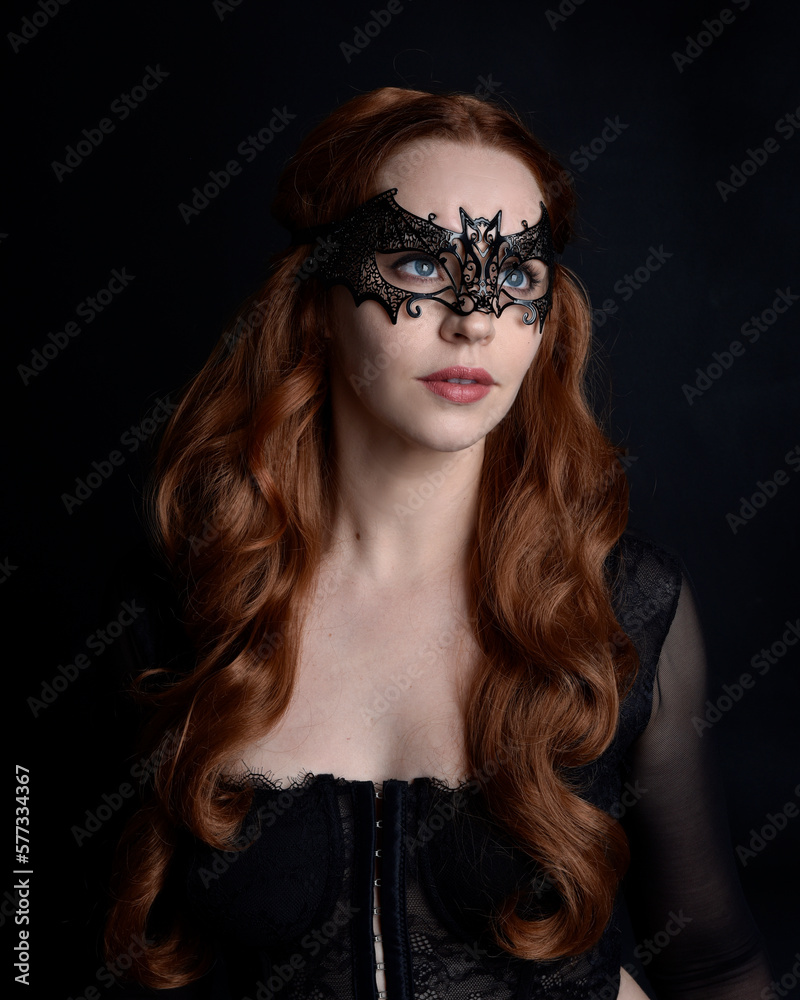 Naklejka premium Masked close up portrait of beautiful woman with long red hair wearing sheer corset top, and black bat wing mask. Isolated on dark studio background with. Moody silhouette lighting.