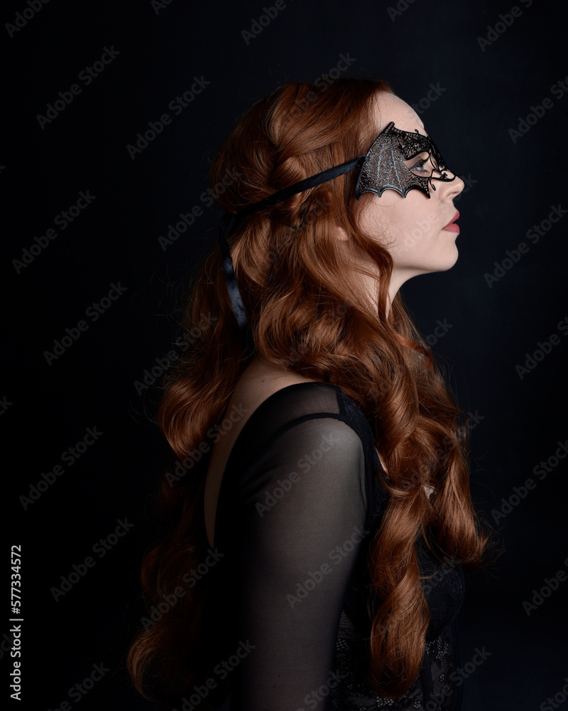 Fototapeta premium Masked close up portrait of beautiful woman with long red hair wearing sheer corset top, and black bat wing mask. Isolated on dark studio background with. Moody silhouette lighting.