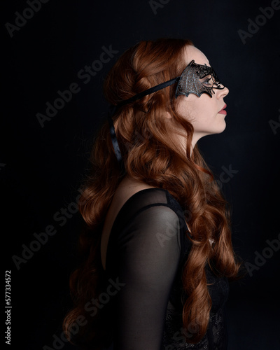 Masked close up portrait of beautiful woman with long red hair wearing sheer corset top  and black bat wing mask.  Isolated on dark studio background with. Moody silhouette lighting.