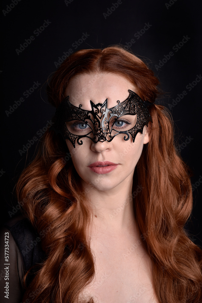 Masked close up portrait of beautiful woman with long red hair wearing sheer corset top, and black bat wing mask. Isolated on dark studio background with. Moody silhouette lighting. Masked close up portrait of beautiful woman with long red hair wearing sheer corset top, and black bat wing mask. Isolated on dark studio background with. Moody silhouette lighting.