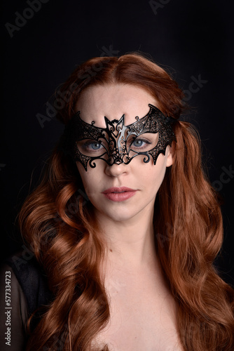 Masked close up portrait of beautiful woman with long red hair wearing sheer corset top, and black bat wing mask. Isolated on dark studio background with. Moody silhouette lighting.