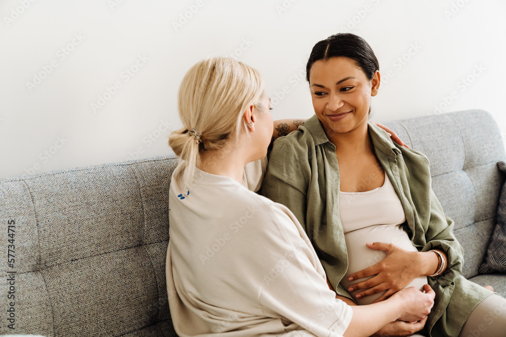Pregnant woman smiling while sitting with her wife on sofa Pregnant woman smiling while sitting with her wife on sofa