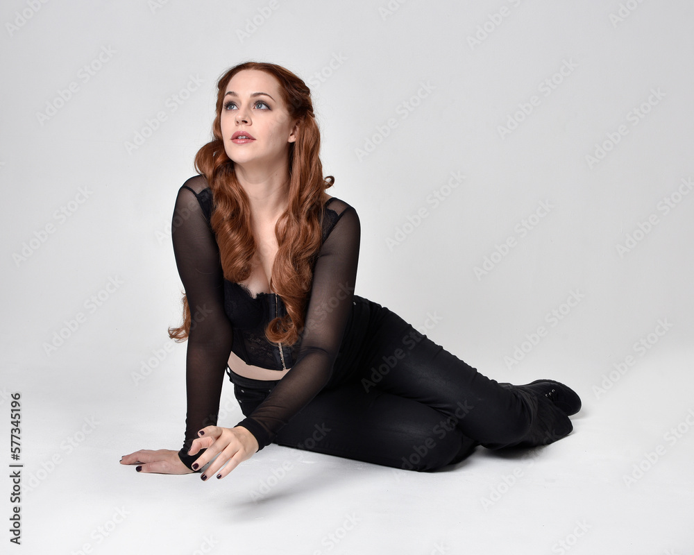 Full length portrait of beautiful woman with long red hair wearing sheer corset top and leather pants. Sitting pose with gestural hands reaching out, kneeling on floor. Isolated on white studio backg Full length portrait of beautiful woman with long red hair wearing sheer corset top and leather pants. Sitting pose with gestural hands reaching out, kneeling on floor. Isolated on white studio backg