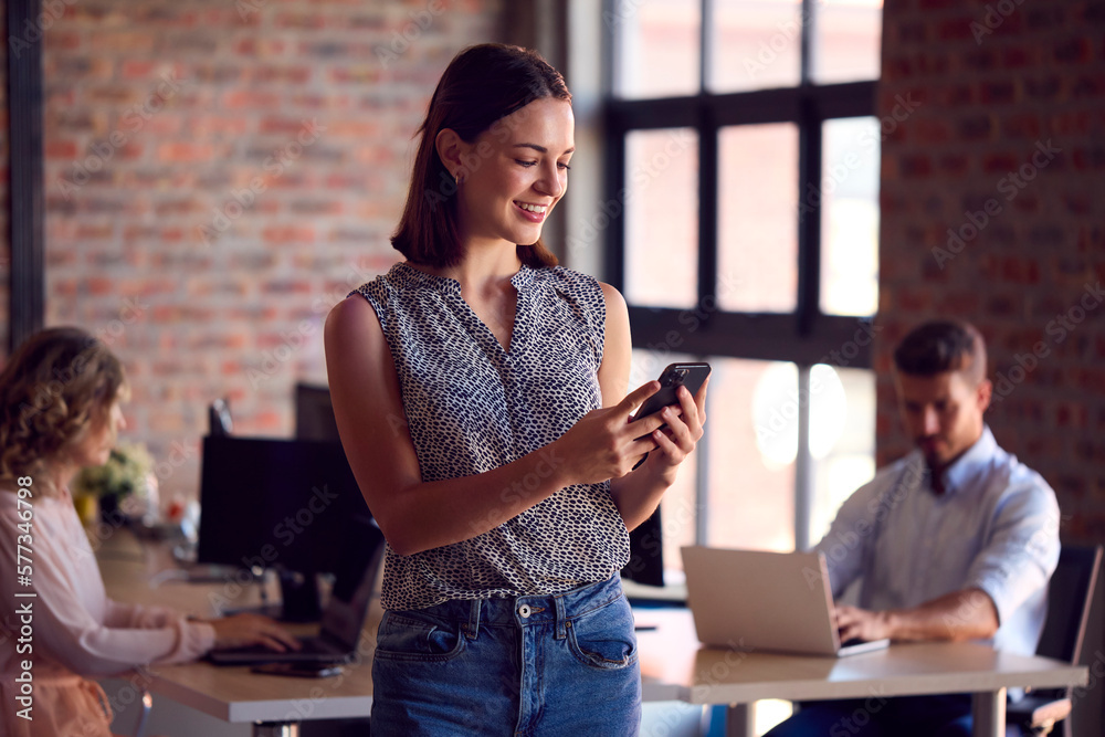 Young Smiling Businesswoman Standing In Busy Office Messaging On Mobile Phone Young Smiling Businesswoman Standing In Busy Office Messaging On Mobile Phone
