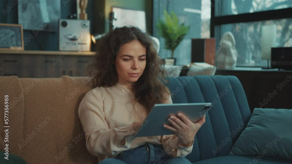 Horizontal footage of attractive young elegant woman with cheerful smile looking directly at camera. Cute smiling girl touching screen of gadget, reading messages indoor. Relax and comfort
