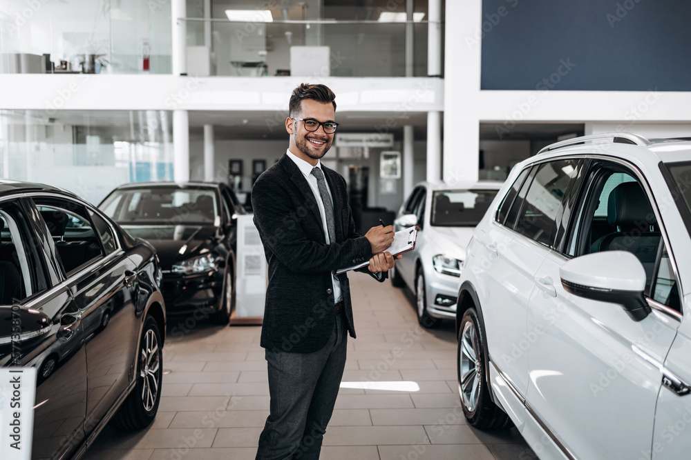 Good looking, cheerful and friendly salesman poses in a car salon or showroom.