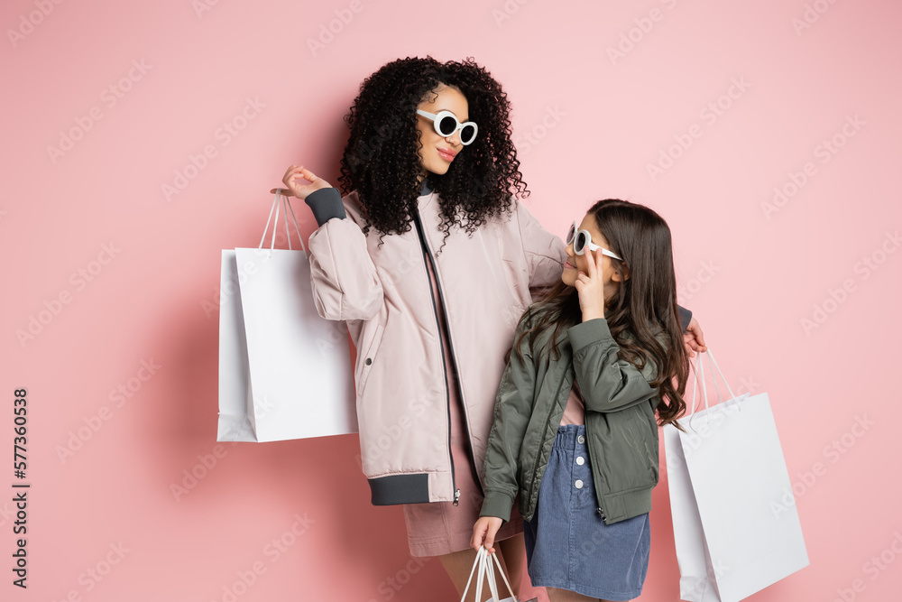 Φωτογραφία Trendy woman and kid in sunglasses holding shopping bags on pink background