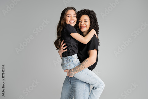 Cheerful tattooed mom holding preteen kid isolated on grey.