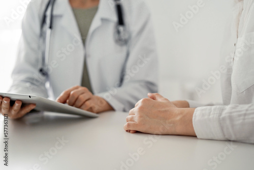Doctor and patient sitting at the desk in clinic office. The focus is on female physician's hands using tablet computer, close up. Perfect medical service and medicine concept.