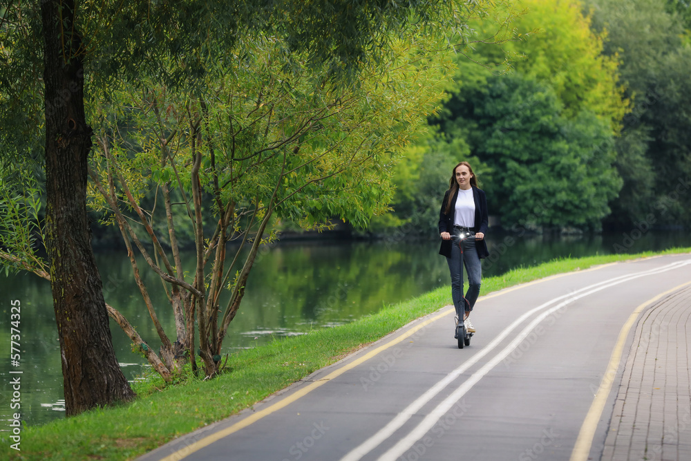 A young woman in stylish clothes rides an electric scooter on a bike path