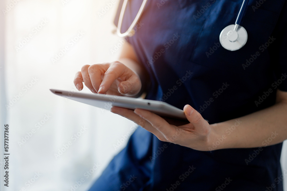 Obraz premium Doctor woman using tablet computer while standing near panorama window in clinic, close up. Physician or surgeon at work. Medicine concept.