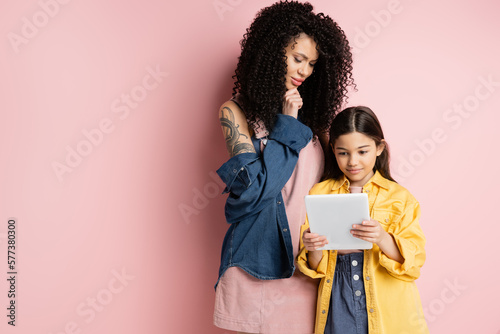Pensive mother looking at digital tablet near daughter on pink background.