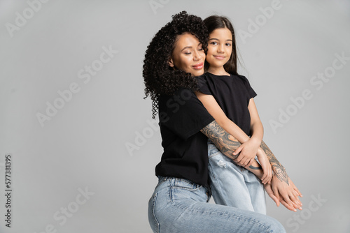Tattooed mother hugging smiling daughter isolated on grey.