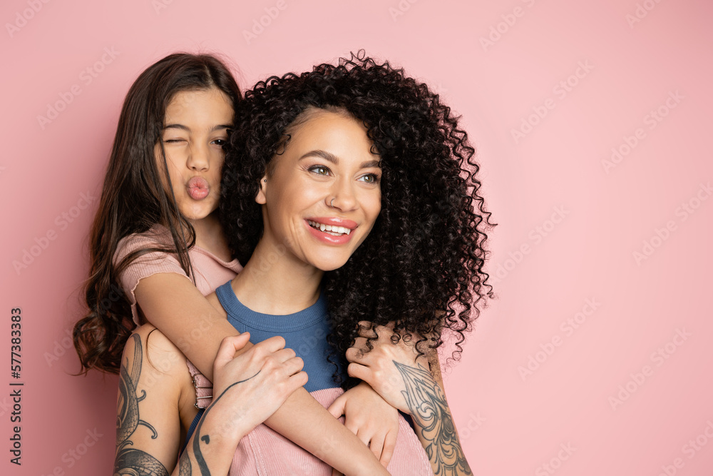 Preteen kid hugging tattooed mom and pouting lips on pink background. Preteen kid hugging tattooed mom and pouting lips on pink background.