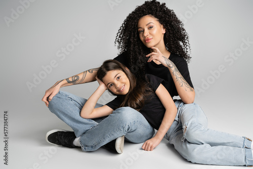 Cheerful kid looking at camera near tattooed mom on grey background.