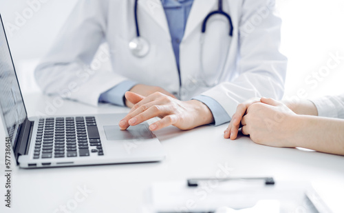Doctor and patient sitting near each other at the desk in clinic. The focus is on female physician s hands using laptop computer  close up. Medicine concept.