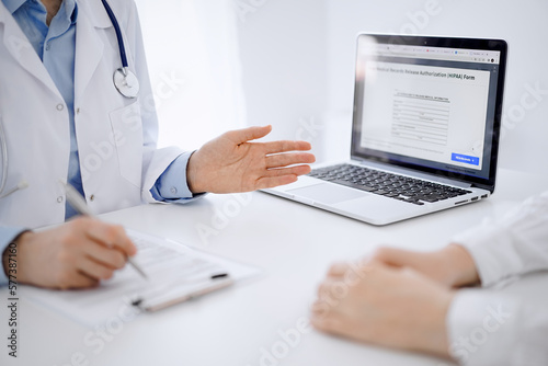 Doctor and patient sitting opposite each other at the desk in clinic. The focus is on female physician s hands pointing into laptop computer monitor  close up. Medicine concept.
