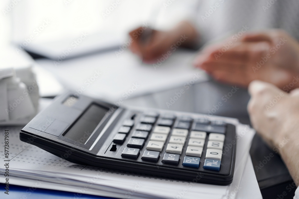 Woman accountant using a pen and laptop computer while counting and discussing taxes with a client, focus on the calculator. Business audit and finance concepts. Woman accountant using a pen and laptop computer while counting and discussing taxes with a client, focus on the calculator. Business audit and finance concepts.