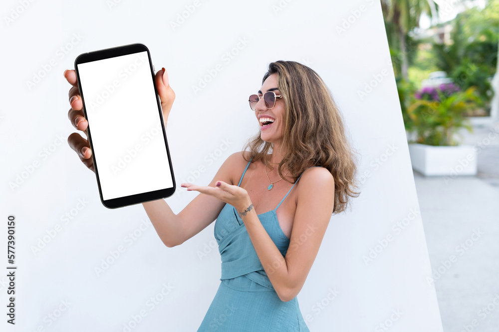 Portrait of beautiful woman in green summer dress on white background natural daylight, pointing on empty white big screen of smartphone, space for advertising