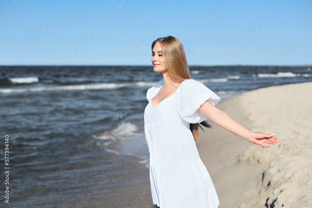 Happy, beautiful woman on the ocean beach standing in a white summer dress, open arms.